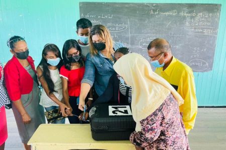 Minister of Education Priya Manickchand cutting the seal off the first box containing the Because We Care Cash Grants at the Cotton Field Secondary School in the company of students, parents, teachers and regional officials. (Ministry of Education photo)