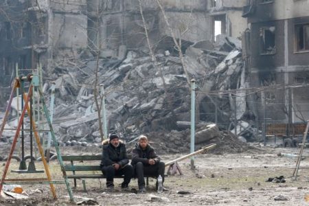 Local residents sit on a bench near an apartment building destroyed in the course of Ukraine-Russia conflict in the besieged southern port city of Mariupol, Ukraine, Mar 25, 2022. (Reuters/Alexander Ermochenko)