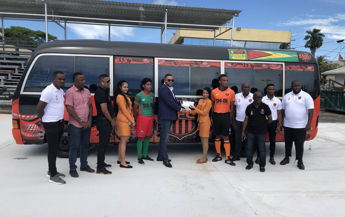 Slingerz FC President Javed Ali collecting a sponsorship cheque from DeAndrea Yansen of newest sponsor Forrester Lumberyard and Building Complex in the presence of other club officials in front of the team bus