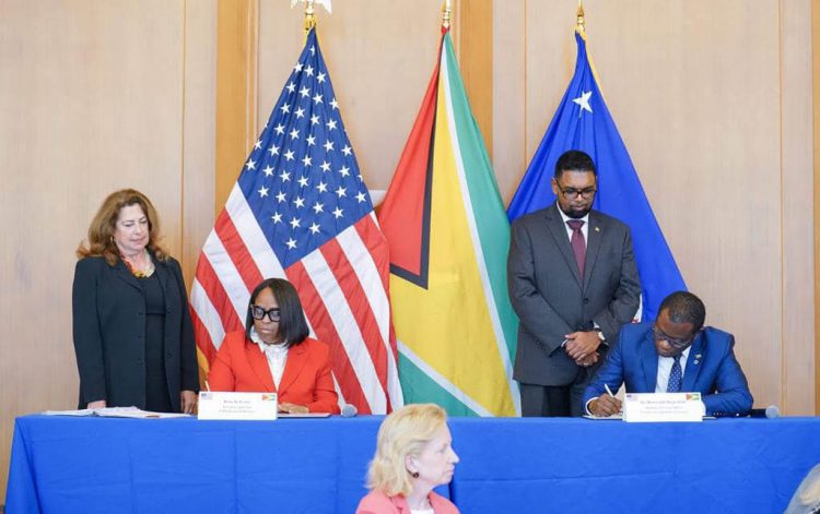 President Irfaan Ali (standing at right) looks on while Minister of Foreign Affairs and Cooperation Hugh Todd (sitting at right) signs the MoU with EXIM Bank’s President Reta Jo Lewis (seated at left). First Vice President and Vice Chair of the Board of Directors at the bank, Judith Pryor is standing at left.   In the foreground is US Ambassador to Guyana Sarah-Ann Lynch. (Office of the President photo) 