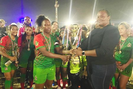 Women’s Football Association President Andrea Johnson presenting the championship trophy to the GDF captain in the presence of teammates following their victory over Fruta Conquerors in the final of the Developmental League