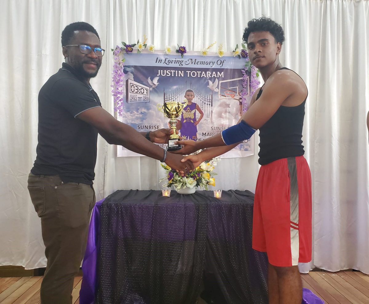 Youth Basketball Guyana’s Co-director Chris Bowman (left) presents the winner’s trophy to Jayden Rampersaud, captain of the winning team in front of a banner honouring the late Justin Totaram