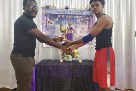 Youth Basketball Guyana’s Co-director Chris Bowman (left) presents the winner’s trophy to Jayden Rampersaud, captain of the winning team in front of a banner honouring the late Justin Totaram